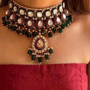 May include: A gold-toned choker necklace featuring square and teardrop-shaped settings with white stones, deep red enamel borders, and dangling dark green beads. The central pendant mirrors the necklace design with additional green bead accents.