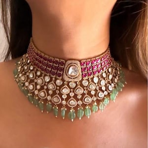 May include: A luxurious choker necklace featuring a central square-shaped diamond surrounded by smaller diamonds. The necklace has a band of red gemstones and dangling green beads, set in a gold-toned metal.