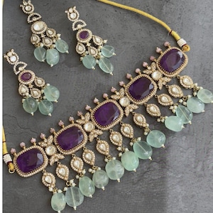 Puede incluir: Un conjunto de joyas de color dorado con un collar y pendientes a juego. El collar y los pendientes tienen grandes piedras preciosas ovaladas de color p&uacute;rpura oscuro, rodeadas de piedras claras m&aacute;s peque&ntilde;as y detalles en forma de l&aacute;grima. Los pendientes y el collar tambi&eacute;n tienen cuentas redondas de color verde claro.
