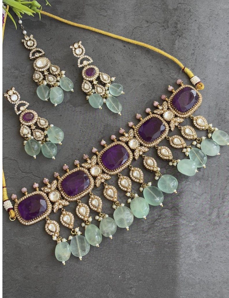 Puede incluir: Un conjunto de joyas de color dorado con un collar y pendientes a juego. El collar y los pendientes tienen grandes piedras preciosas ovaladas de color p&uacute;rpura oscuro, rodeadas de piedras claras m&aacute;s peque&ntilde;as y detalles en forma de l&aacute;grima. Los pendientes y el collar tambi&eacute;n tienen cuentas redondas de color verde claro.