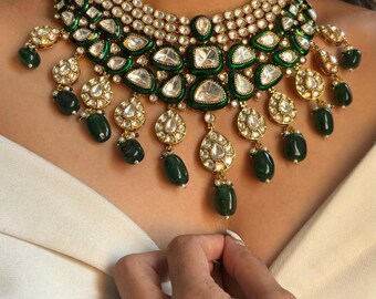 May include: A gold-toned necklace with emerald green and clear gemstone accents. The necklace features multiple layers of stones and dangling emerald green teardrop-shaped gems. A silver ring is visible on a finger.