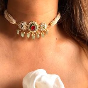 May include: A gold choker necklace featuring a central red gemstone, surrounded by white and green stones. The necklace has a braided, off-white cord and dangling green teardrop accents. The necklace is worn around the neck.
