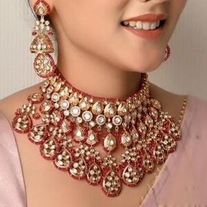Könnte beinhalten: Eine gold-rote Halskette und passende Ohrringe. Die Halskette ist im Choker-Stil mit aufwendigen Details und tropfenförmigen Anhängern. Die Ohrringe sind im Kronleuchter-Stil mit ähnlichen Designelementen. Der Schmuck ist vor einem rosa Hintergrund platziert.