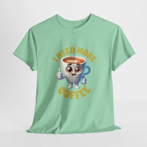 Op de afbeelding: Een lichtgroen T-shirt met een cartoon koffiekopje met een gezicht en de tekst "Ik heb meer koffie nodig" in geel.