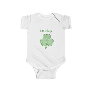 以下が含まれることがあります： 白いベビーボディスーツ。緑色の「Lucky」の文字と、緑色のクローバーのイラストが描かれています。クローバーはハート型の葉が3枚と茎があります。ボディスーツは半袖で、スナップ留めです。