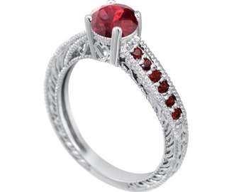 May include: A silver-toned ring featuring a large, round, faceted red center stone held by four prongs. The band is decorated with intricate filigree metalwork and a row of five small, round, red accent stones set along each side of the shank.
