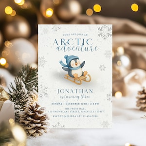 May include: A light blue and white invitation card with the text "Arctic Adventure". It features a cartoon penguin on a sled, wearing a blue hat and scarf. The card also includes event details: "Jonathan is turning three".