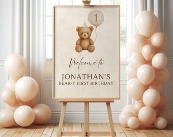 May include: A wooden easel displays a beige sign with a teddy bear illustration and the text "Welcome to Jonathan's Bear-y First Birthday." A balloon with the number "1" floats above the bear. Peach-colored balloons are arranged around the easel.