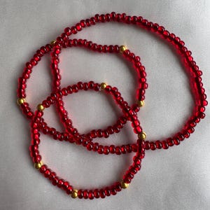 Könnte beinhalten: Drei rote Perlenarmbänder mit goldenen Akzenten. Die Armbänder sind übereinander gelegt.
