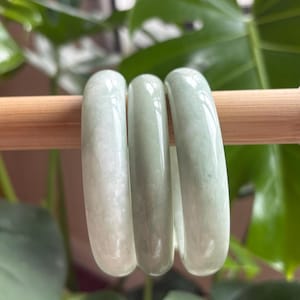 Puede incluir: Tres pulseras de jade verde apiladas sobre una superficie de madera. Las pulseras son lisas y pulidas.