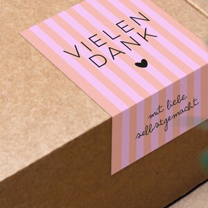 Puede incluir: Una etiqueta de regalo a rayas rosa y blancas con el texto "VIELEN DANK" y un pequeño corazón. La etiqueta también dice "mit liebe selbstgemacht", que se traduce como "hecho con amor".
