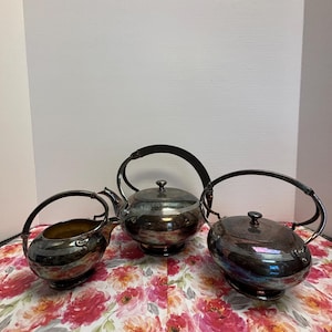 May include: A silver tea set comprising a teapot, a creamer, and a sugar bowl. The teapot features a tall handle and a lid. The creamer and sugar bowl have smaller handles. The set is displayed on a floral tablecloth.