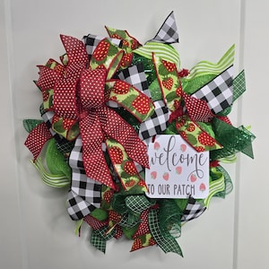 May include: A decorative wreath featuring red and white polka dot bows, black and white checkered ribbons, and green mesh. The wreath includes ribbons with strawberry patterns and a small sign that reads "welcome to our patch."