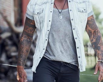 May include: A man wearing a white denim shirt with short sleeves, a grey t-shirt, black jeans, and a silver necklace with a dog tag pendant. He has tattoos on both arms.