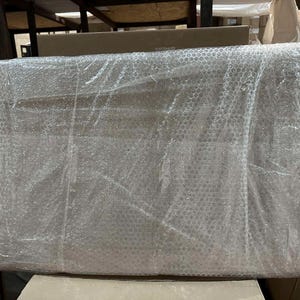 May include: A rectangular object wrapped in clear bubble wrap. The object is large and appears to be a piece of furniture or a similar item, ready for shipping. The bubble wrap provides protection during transit.