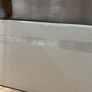 May include: A large, rectangular piece of corrugated cardboard, likely for packaging or shipping. The cardboard is a light tan color with visible horizontal ridges. The image is taken from a slightly angled perspective, showing the top and side of the cardboard.