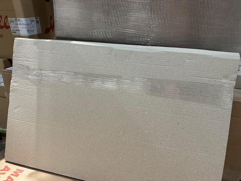 May include: A large, rectangular piece of corrugated cardboard, likely for packaging or shipping. The cardboard is a light tan color with visible horizontal ridges. The image is taken from a slightly angled perspective, showing the top and side of the cardboard.