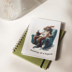 May include: A white journal with a cartoon dragon wearing a "Best Dad" t-shirt, relaxing in a blue armchair, holding a remote. The journal is stacked on a green spiral notebook and a green notebook with an elastic band. The text "musings of a legend" is below the dragon.