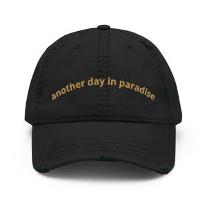 Op de afbeelding: Zwarte, distressed baseballcap met de gouden geborduurde tekst "another day in paradise". De cap heeft een gebogen klep en een versleten, vintage uitstraling.