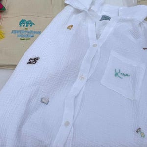 May include: White button-down shirt with small embroidered designs and a pocket with the name "Kara" in green. A canvas tote bag with the text "The Adventurous Nomads Travel Club" is visible in the background.