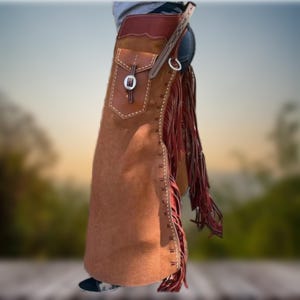 May include: Brown leather chaps with fringe and a decorative pocket. The chaps have a red stitching detail and a silver buckle.