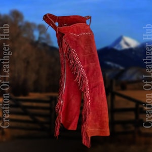 May include: Red suede chaps with fringe and white stitching. The chaps are hanging on a fence with a mountain range in the background.