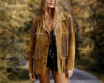 May include: A brown suede jacket with fringe and gold studs. The jacket has a button-up front and long sleeves. The model is wearing a white beanie and has long blonde hair.