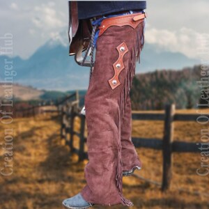 May include: Brown suede chaps with fringe and a leather belt with silver accents. The chaps are worn by a person standing in a field with a wooden fence in the background.
