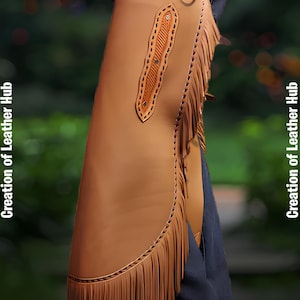 May include: A brown leather chaps with fringe detail and a decorative stitching pattern. The chaps are worn over blue jeans and have a belt loop at the top.