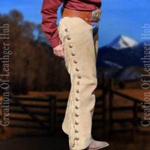 May include: A pair of beige leather chaps with fringe and silver snaps. The chaps are worn by a person standing in front of a wooden fence and a mountain range in the background.