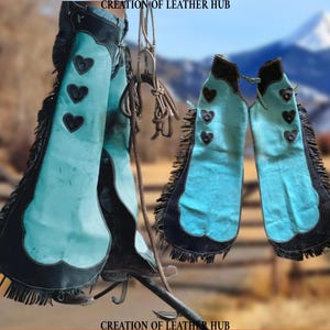 May include: A pair of turquoise leather chaps with black fringe and heart-shaped cutouts. The chaps are hanging on a fence post with a mountain range in the background.