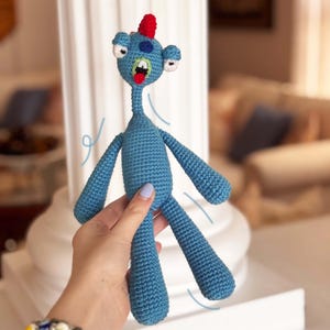 May include: A blue crocheted monster toy with a red and white spiky hair, three eyes, and a wide open mouth. The toy is being held by a hand.