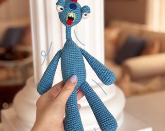 May include: A blue crocheted monster toy with a red and white spiky hair, three eyes, and a wide open mouth. The toy is being held by a hand.
