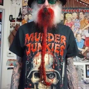 Könnte beinhalten: Schwarzes T-Shirt mit den Worten "MURDER JUNKIES" in roten, tropfenden Buchstaben. Das Shirt zeigt einen Schädel mit Helm und Messer. Die Person trägt einen schwarzen Hut und grüne Sonnenbrille.