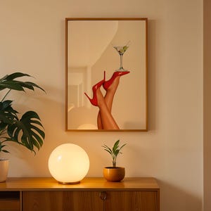 Framed print hung on a neutral wall with a glowing orb lamp and monstera plant nearby. The red heels pop against the warm evening ambiance.