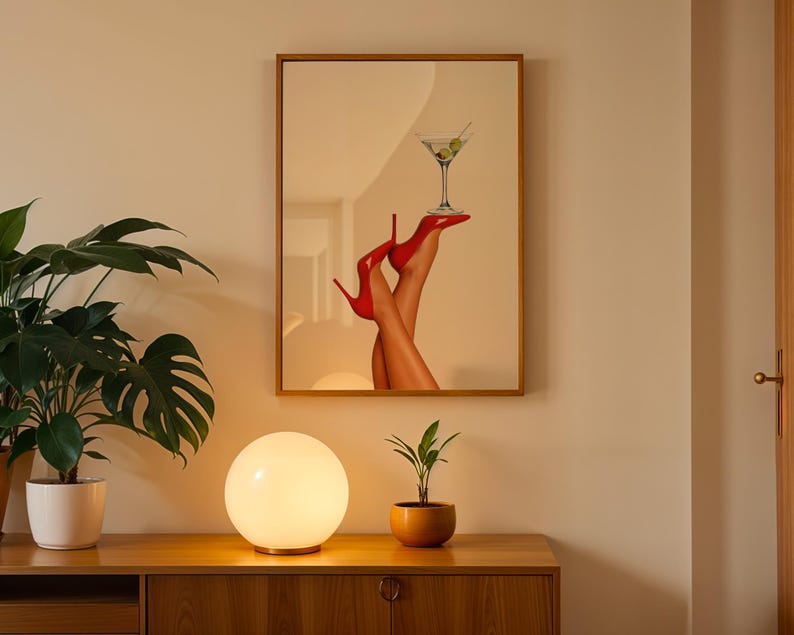 Framed print hung on a neutral wall with a glowing orb lamp and monstera plant nearby. The red heels pop against the warm evening ambiance.