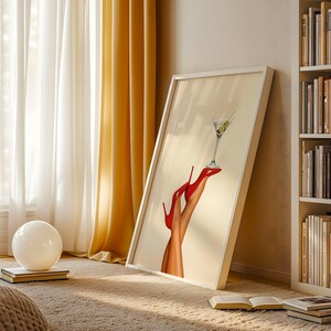 Artwork leaning against a bookcase in a cozy, golden-lit room. The red heels and martini glass catch sunlight on a textured beige blanket, adding glamour to the relaxed space.