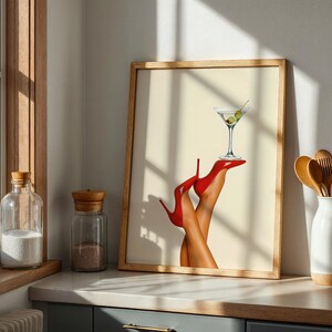 Framed print leaning against a wall in a modern kitchen. Sunlight highlights the red heels and martini glass, adding flair to the neutral counter setup.