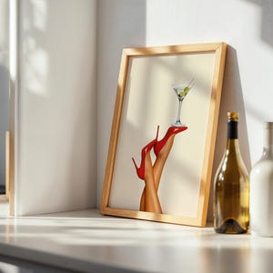 Framed print on a sunlit white table beside a wine bottle and glass. The red heels and martini glass are bright and elegant in a minimal dining setup.
