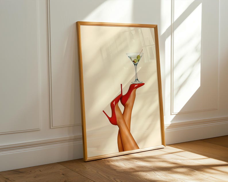 Framed art print of a hand in red high heels holding a martini glass with olives. Leaning against a white wall in soft sunlight on a wooden floor, creating a bold and playful vibe.