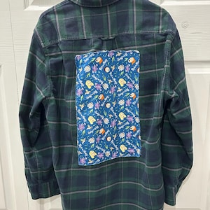 May include: A green and navy plaid flannel shirt with a blue patch featuring cartoon characters and the text "Rick and Morty." The shirt is hanging on a black hanger against a white door.