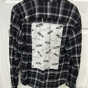 May include: Black and white plaid flannel shirt with long sleeves. A white patch on the back features the text "AC/DC" in a repeating pattern. The shirt is hanging on a black hanger.