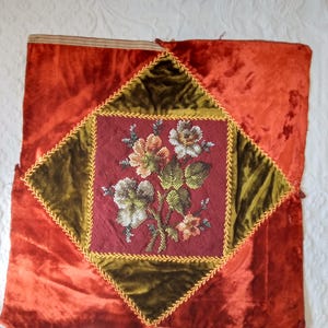 May include: A square, antique textile with a central floral embroidery. The embroidery is framed by a dark green velvet diamond, outlined with gold trim, set on a red velvet background. The floral design features white, orange, and green flowers.