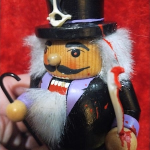 May include: A wooden nutcracker figurine with a black top hat, black coat, and purple accents. The figurine has a white fur collar and a mustache. It holds a cane and a ball, both with red paint dripping down, suggesting a theme of horror.