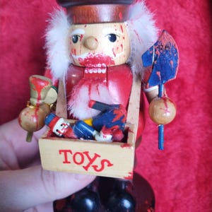 May include: A wooden nutcracker figurine with a red and white uniform, a brown hat, and a white beard. The nutcracker is holding a blue and red axe and a box of toys. The word "TOYS" is printed on the box. The figurine has red paint splatters.