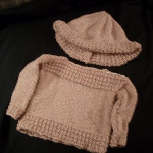 May include: A pink knitted baby sweater and hat set. The sweater has long sleeves and a boat neck. The hat is a simple beanie style.