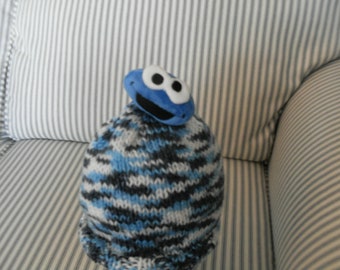 May include: A blue and white knitted hat with a blue cookie monster character on top. The hat is sitting on a striped couch.