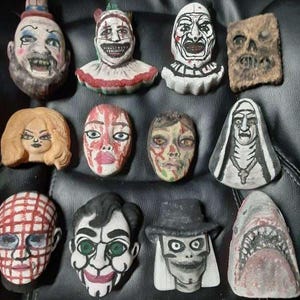 May include: A collection of eleven hand-painted, horror-themed character figurines. The figurines depict various faces, including clowns, a nun, and a shark, with detailed facial features and vibrant colors. The collection is arranged on a black surface.
