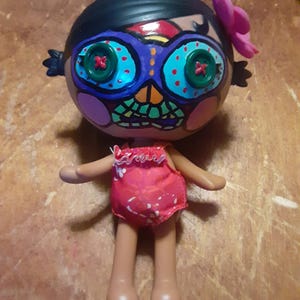 May include: A doll with a painted face in the style of a sugar skull. The face is painted with blue, orange, and green accents, with large button eyes. The doll has a pink flower in its hair and is wearing a red swimsuit.