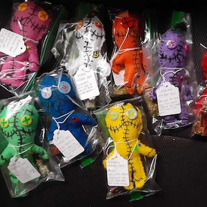 May include: A collection of colorful voodoo dolls with stitched details and button eyes. Each doll is packaged in a clear plastic bag with a tag that describes the doll's purpose. The tags include phrases like "Pink Voodoo Doll", "Love Voodoo Doll", and "Witchcraft Voodoo Doll".